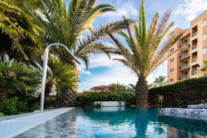 Piscina com deck de azulejos, cercada por palmeiras e um edifício ao fundo.