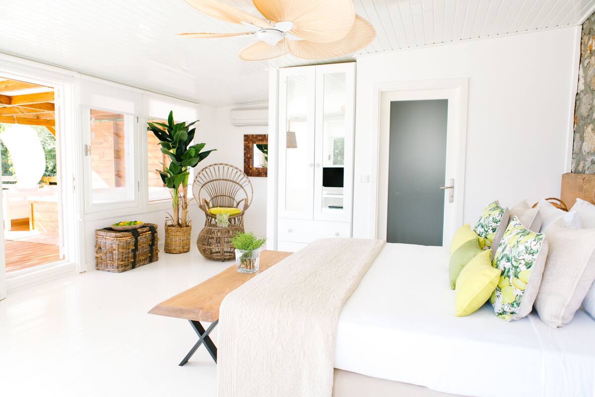 Ein helles, weißes Zimmer mit einem Bett, einem Holztisch, Korbwannen und einem Deckenventilator.