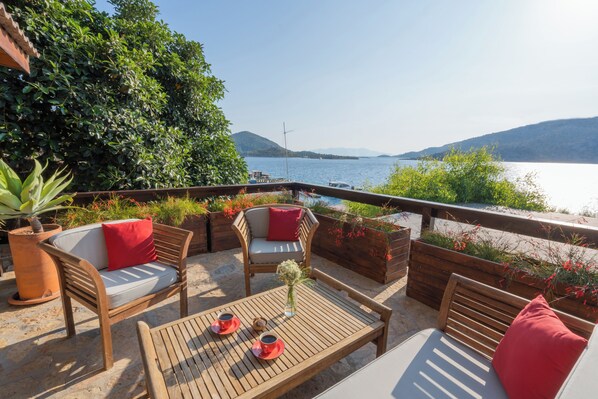 Un espacio al aire libre con mobiliario de madera, una mesa con tazas y vistas a un lago y montañas.