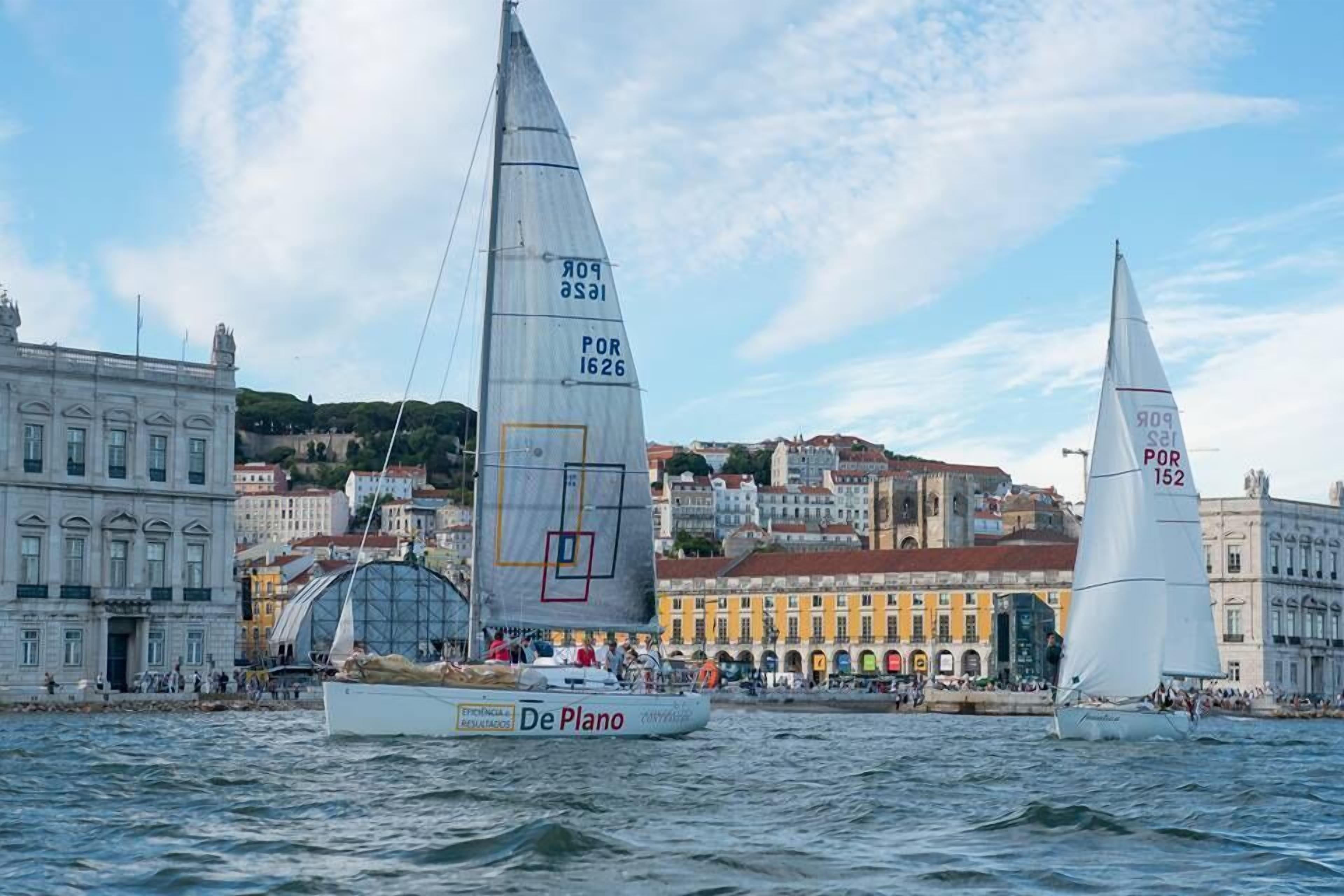 A sailboat named 'De Plano' on a body of water with buildings in the background.