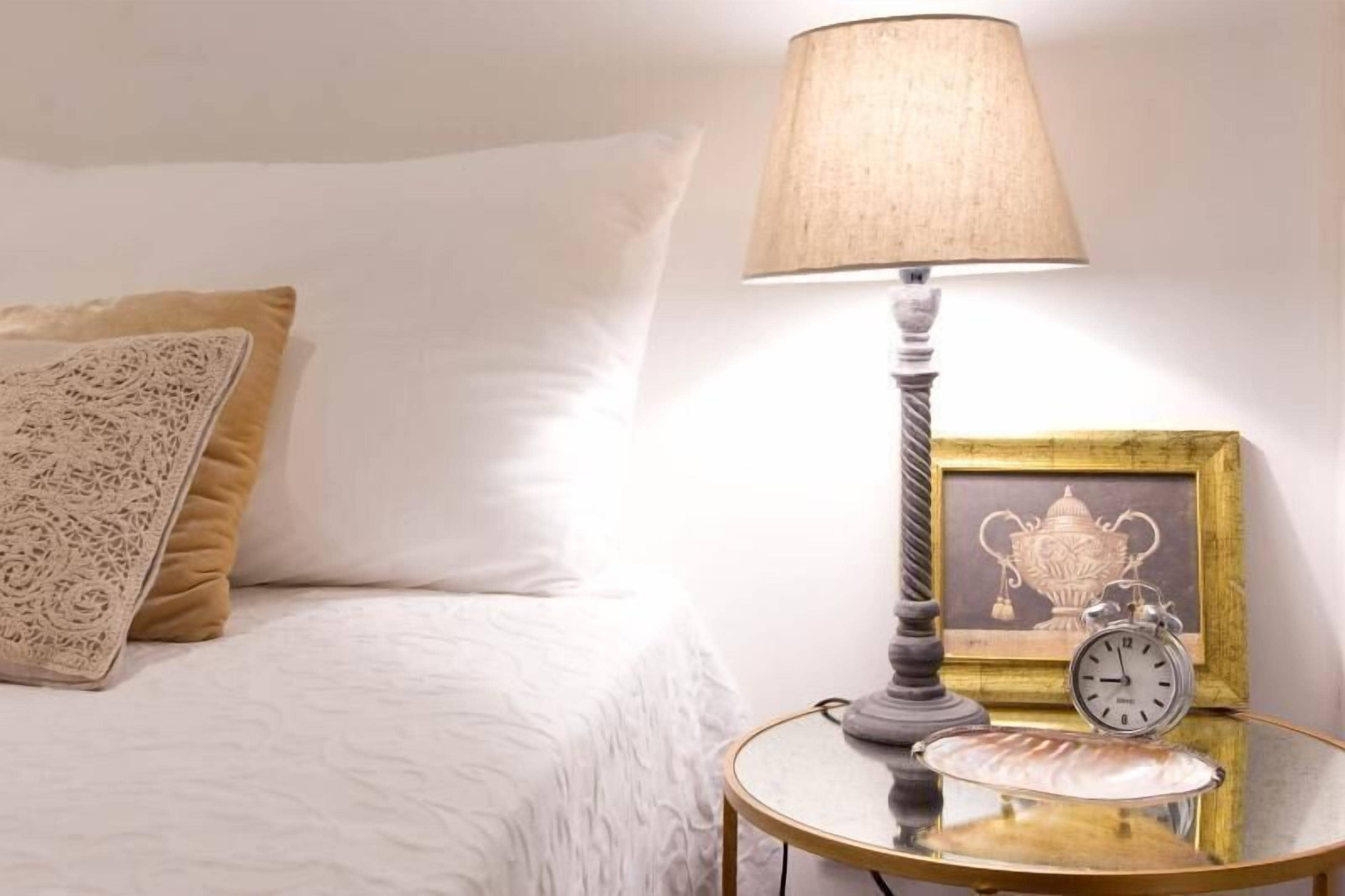 A neatly made bed with a white comforter and a decorative pillow. A table beside the bed holds a lamp, a framed picture, and a small clock.