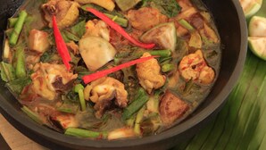 Uma tigela de sopa com legumes e carne, decorada com pimentas vermelhas, servida sobre uma mesa de madeira com uma folha verde e um ramo de flores vermelhas ao fundo.
