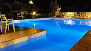Une piscine avec un toboggan, entourée de chaises longues et de tables, et éclairée par un éclairage d’ambiance.