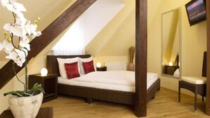 A bedroom with a sloped ceiling, a bed with white bedding and red pillows, a nightstand with a lamp, a potted orchid, a mirror, and a television on the wall. - GuestHouse Heidelberg (Heidelberg)