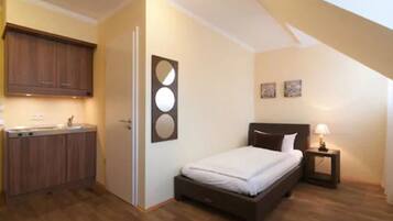 Un dormitorio compacto con una cama individual, una mesita de noche con lámpara y una pequeña kitchenette con armarios de madera.