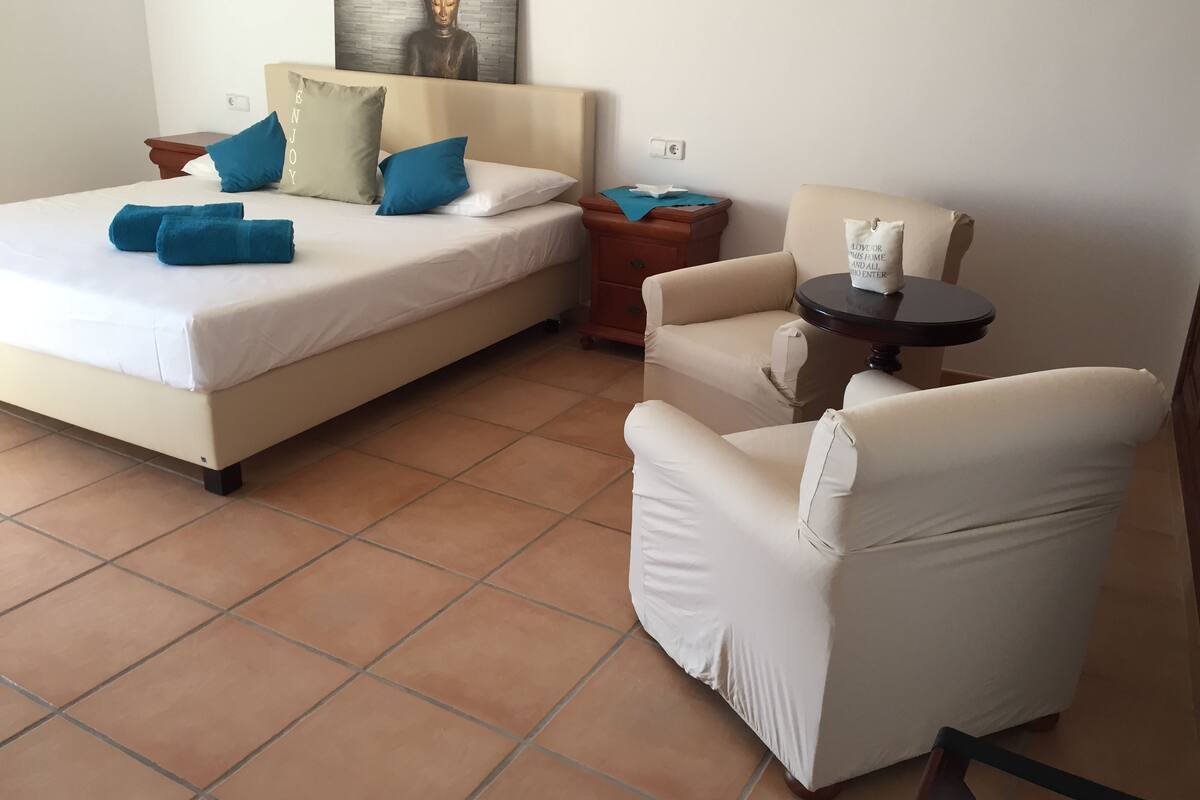 A bedroom with a bed, a chair, a small table, and a Buddha statue on the wall.