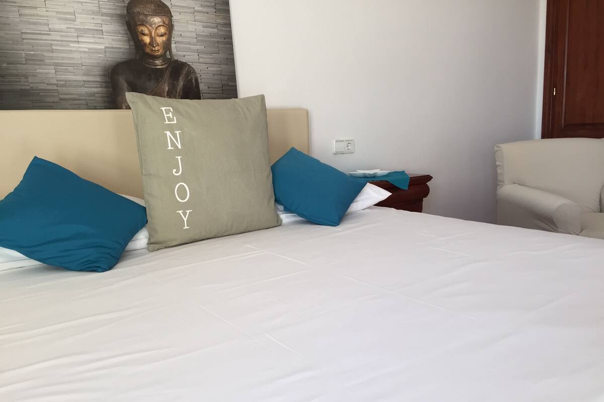A bed with white bedding and teal pillows, a Buddha statue on the wall, and a wooden nightstand with a lamp.