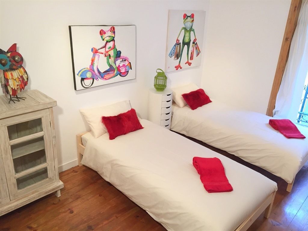 A room with two beds, each with a red pillow and a red towel. There are two colorful paintings on the wall, one of a scooter and one of a frog. A white cabinet with glass doors is visible on the left side.