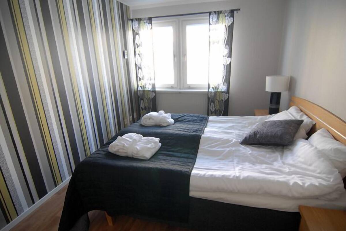 A hotel room with two beds, striped curtains, a window with floral curtains, a bedside lamp, and a wooden headboard.
