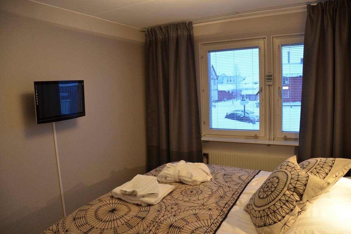 A hotel room with a bed, a flat-screen TV mounted on the wall, and a window with curtains.