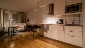 A modern kitchen with white cabinets, a stainless steel range hood, and a built-in oven. There is a bar stool and a small table with a lamp. - Simloc Hotel Drottninggatan (Arjeplog)
