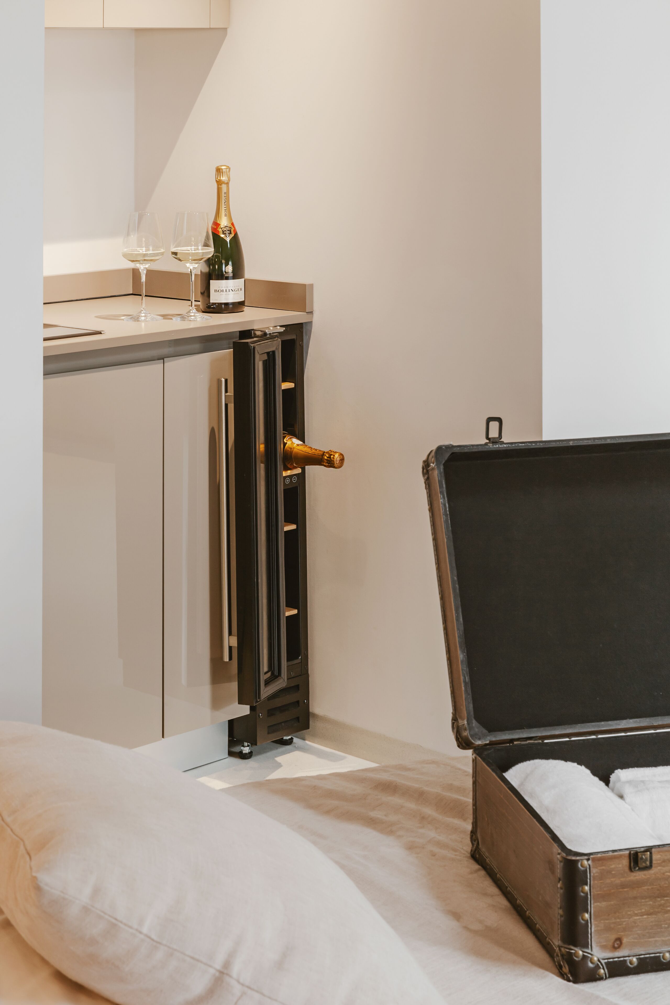 A room with a bed, a chest of drawers, a suitcase, and a bottle of champagne.