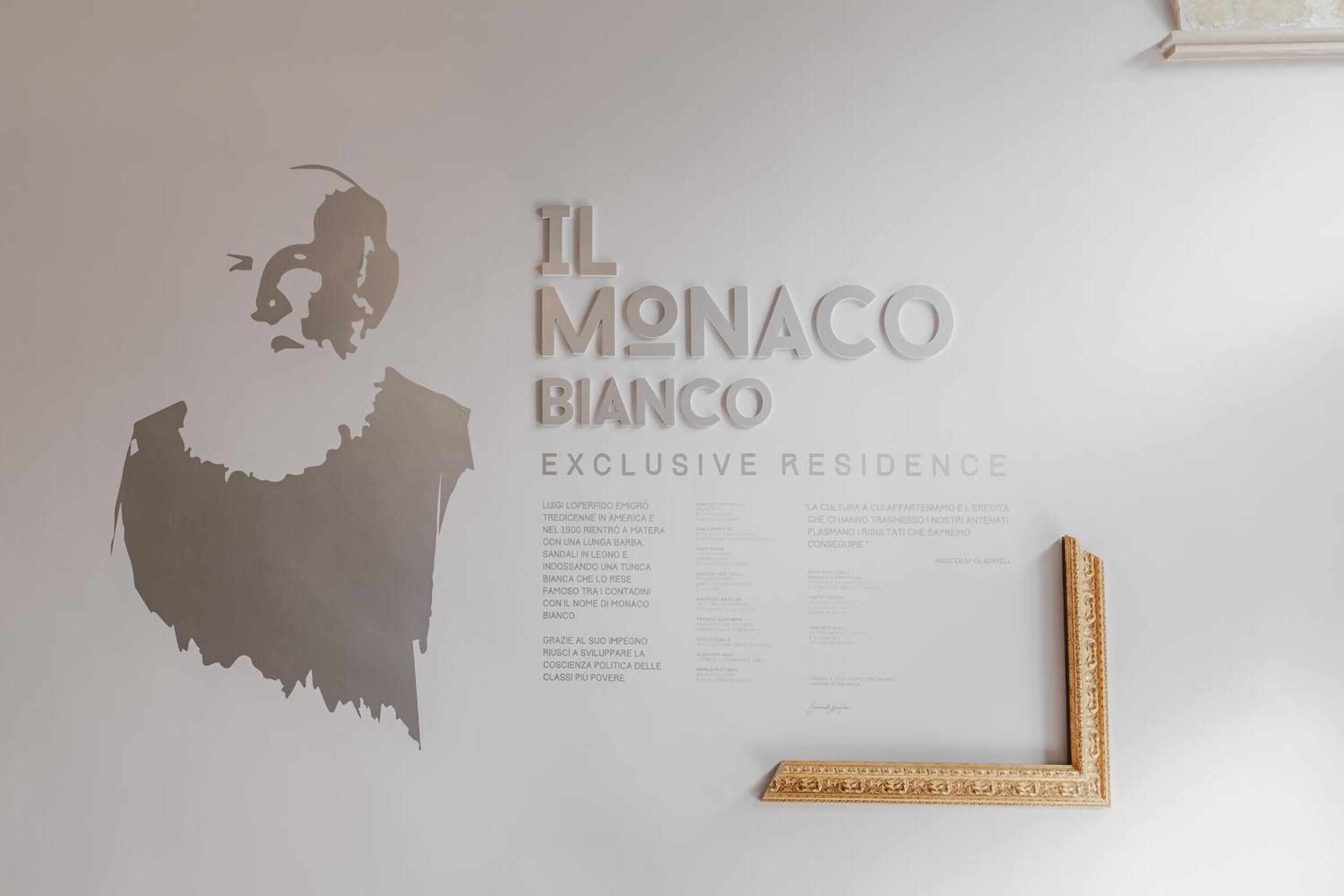 Un mur sur lequel on peut lire l’inscription « IL MONACO BIANCO, résidence de luxe » et où est accrochée une image encadrée.