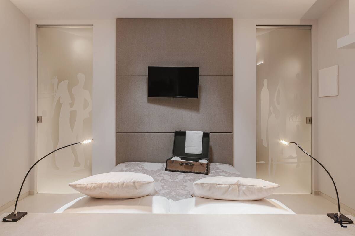 A modern bedroom with a bed, a flat-screen TV, a bedside table, and two floor lamps.