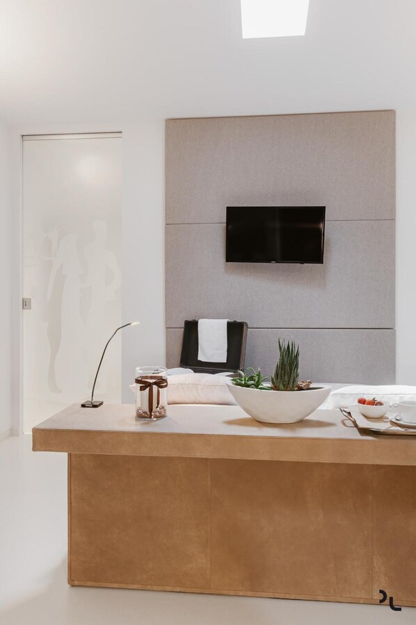 A modern hotel room with a minimalist design, featuring a large window, a flat-screen TV mounted on a grey panel, a wooden bedside table with a vase and a plant, and a white bed with a black towel.