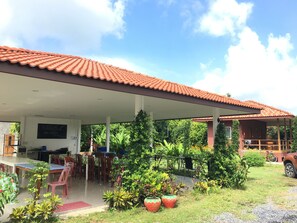 A covered outdoor seating area with tables and chairs, surrounded by potted plants and a red-tiled roof. - Ban Suan Kru Kob (Pak Phli)