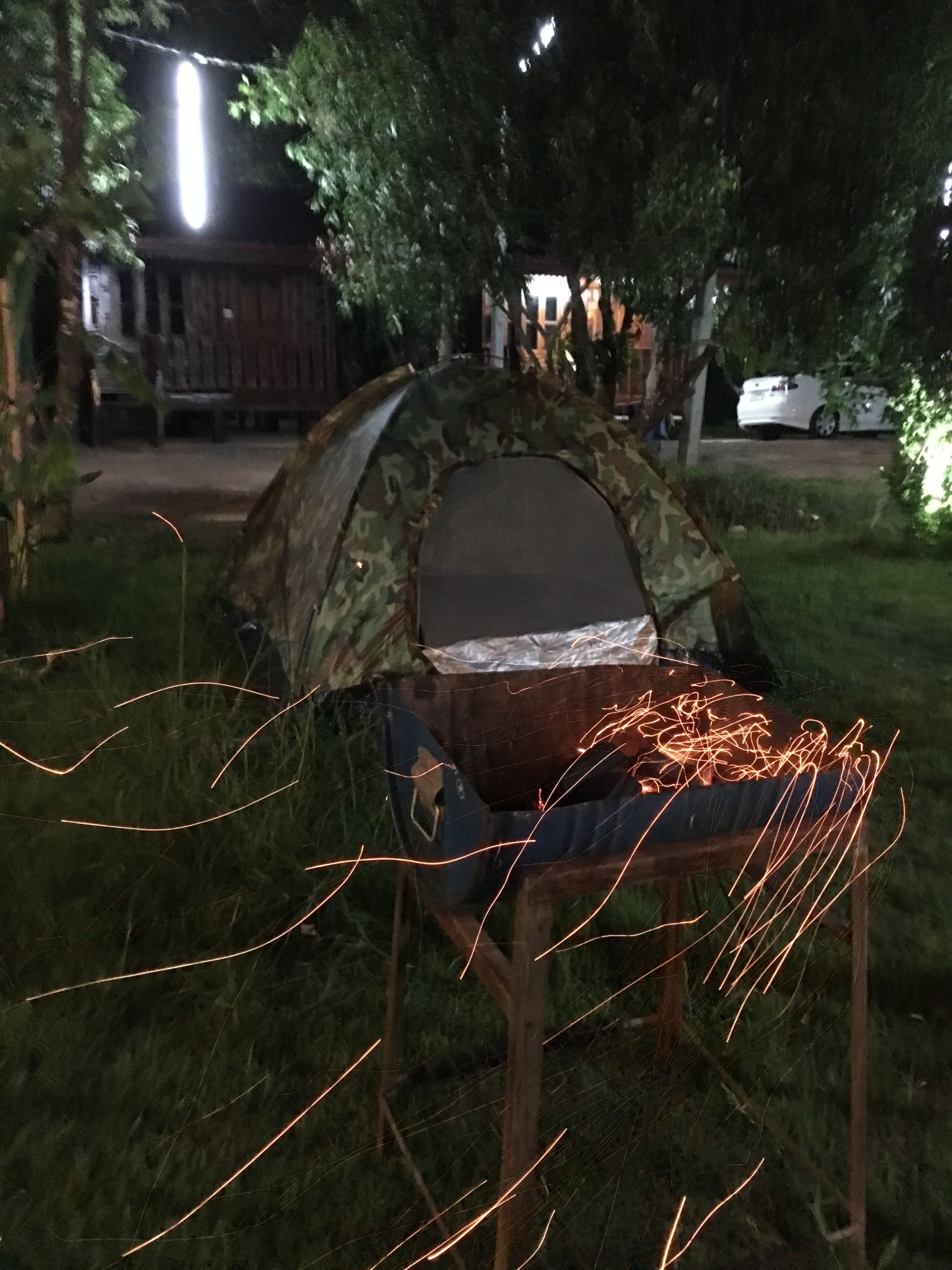 Uma tenda com exterior camuflado e estrutura metálica, montada em área gramada com árvores e um edifício ao fundo.