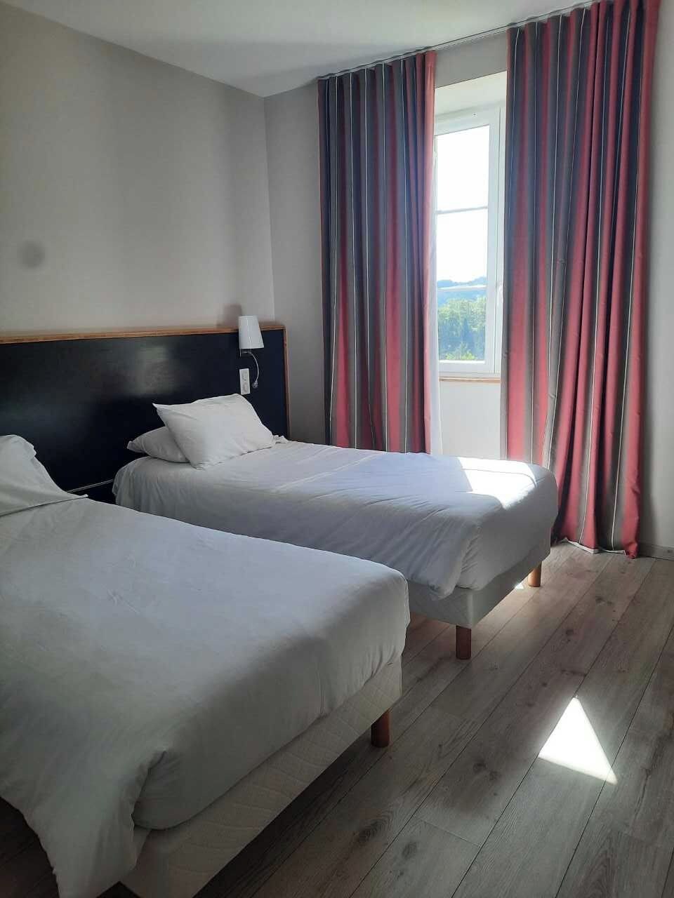 Habitación de hotel con dos camas, una mesita de noche, una lámpara y una ventana con cortinas.