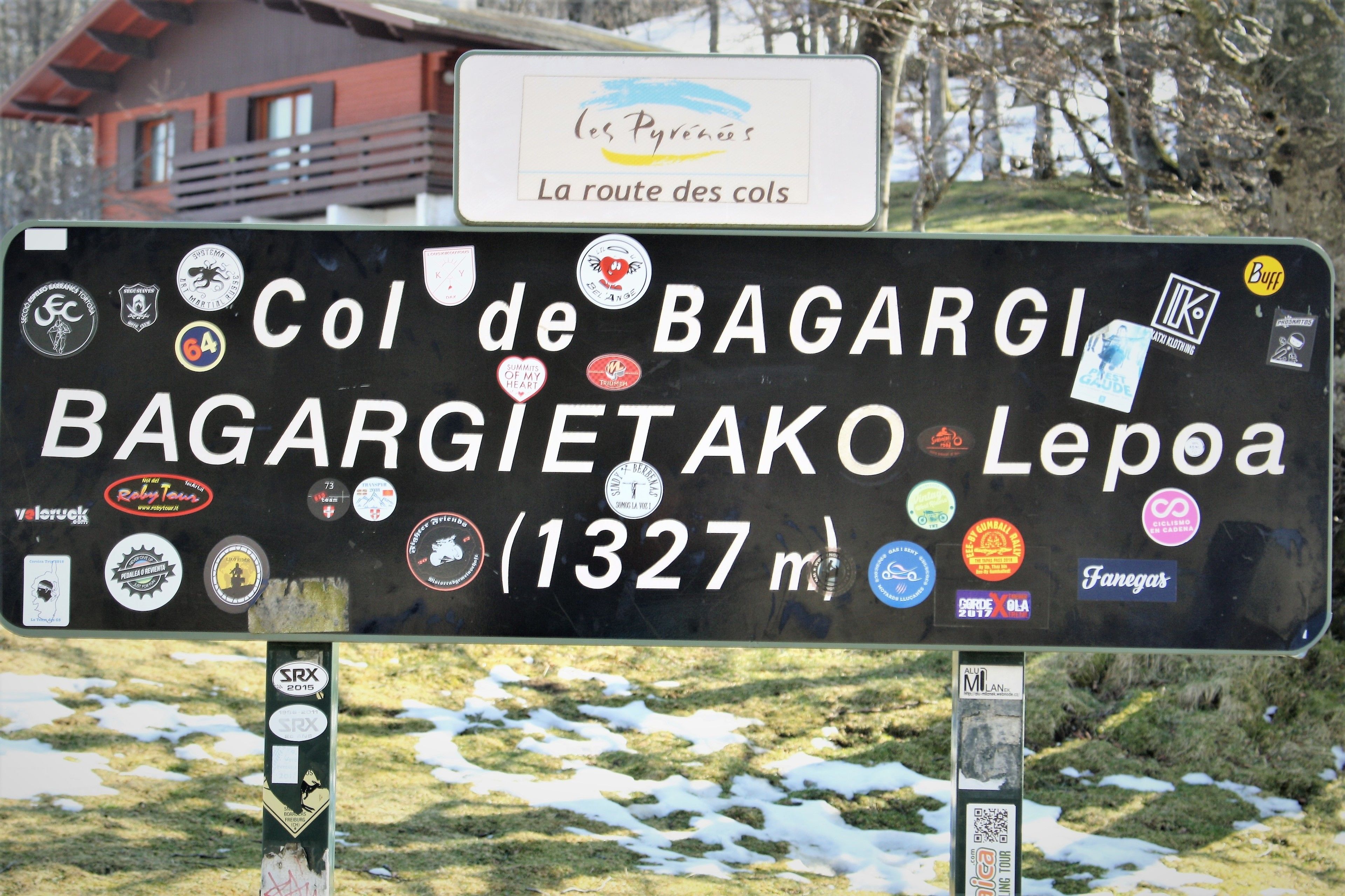 A sign with multiple stickers and text indicating 'Col de BAGARGIETAKO Lepaia' and '1327 m'.