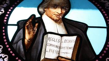 Una vetrata istoriata raffigura una figura che tiene un libro intitolato “REGLES des ECOLES COMMUNESCHRETIENNES des FRERES”.