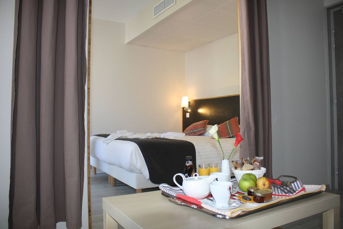 Une chambre d’hôtel avec un lit parfaitement fait, un plateau de petit-déjeuner avec des œufs, des toasts et du jus, et un vase contenant une seule fleur.