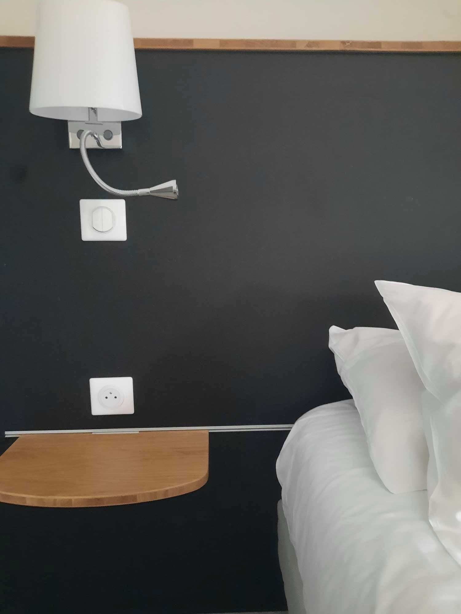 A hotel room with a bed, a nightstand, a lamp, and a power outlet.
