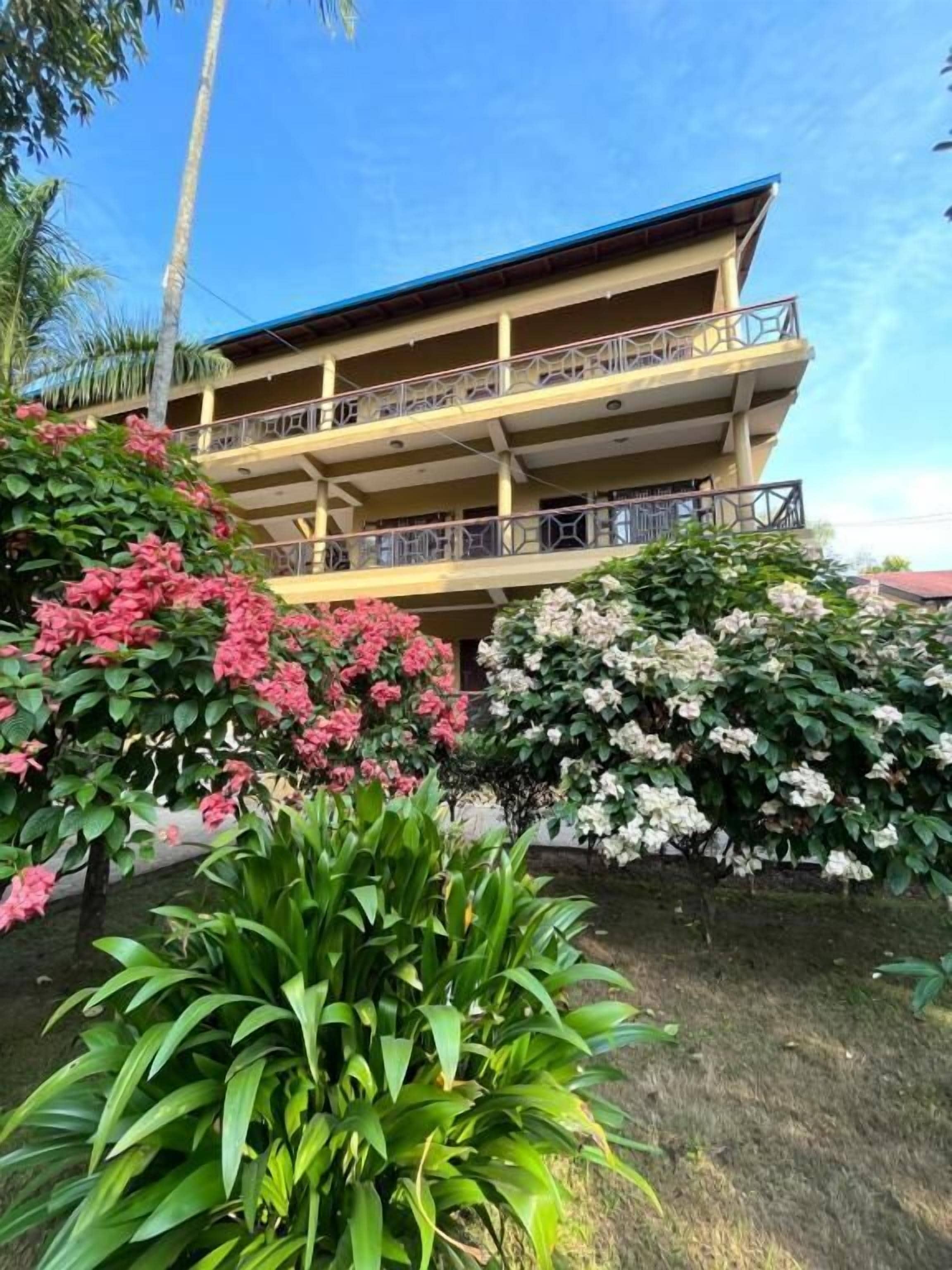 A multi-story building with balconies, surrounded by lush greenery and flowering plants.