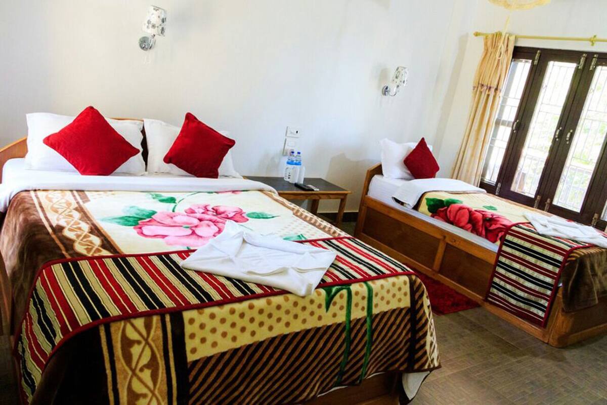 A hotel room with two beds, each with a striped bedspread and red pillows. There is a small table with a bottle of water and a remote control.