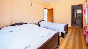 Habitación de hotel con dos camas, ventilador de techo, baño con puerta de vidrio ajedrezada y suelo de madera.