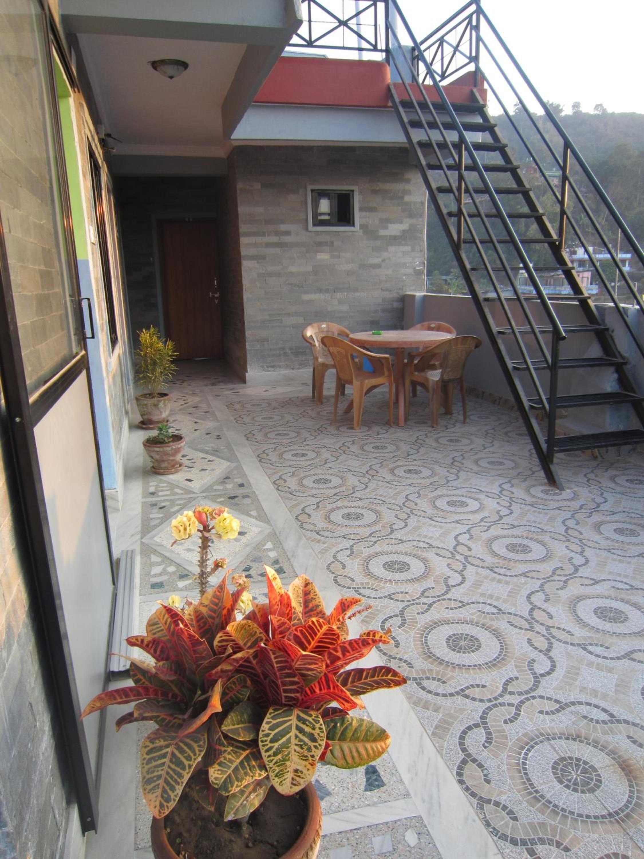 Un patio techado con piso de baldosas, una mesa y sillas de madera, una planta en maceta y una escalera que sube a un nivel superior.