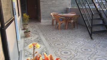 Un patio techado con piso de baldosas, una mesa y sillas de madera, una planta en maceta y una escalera que sube a un nivel superior.