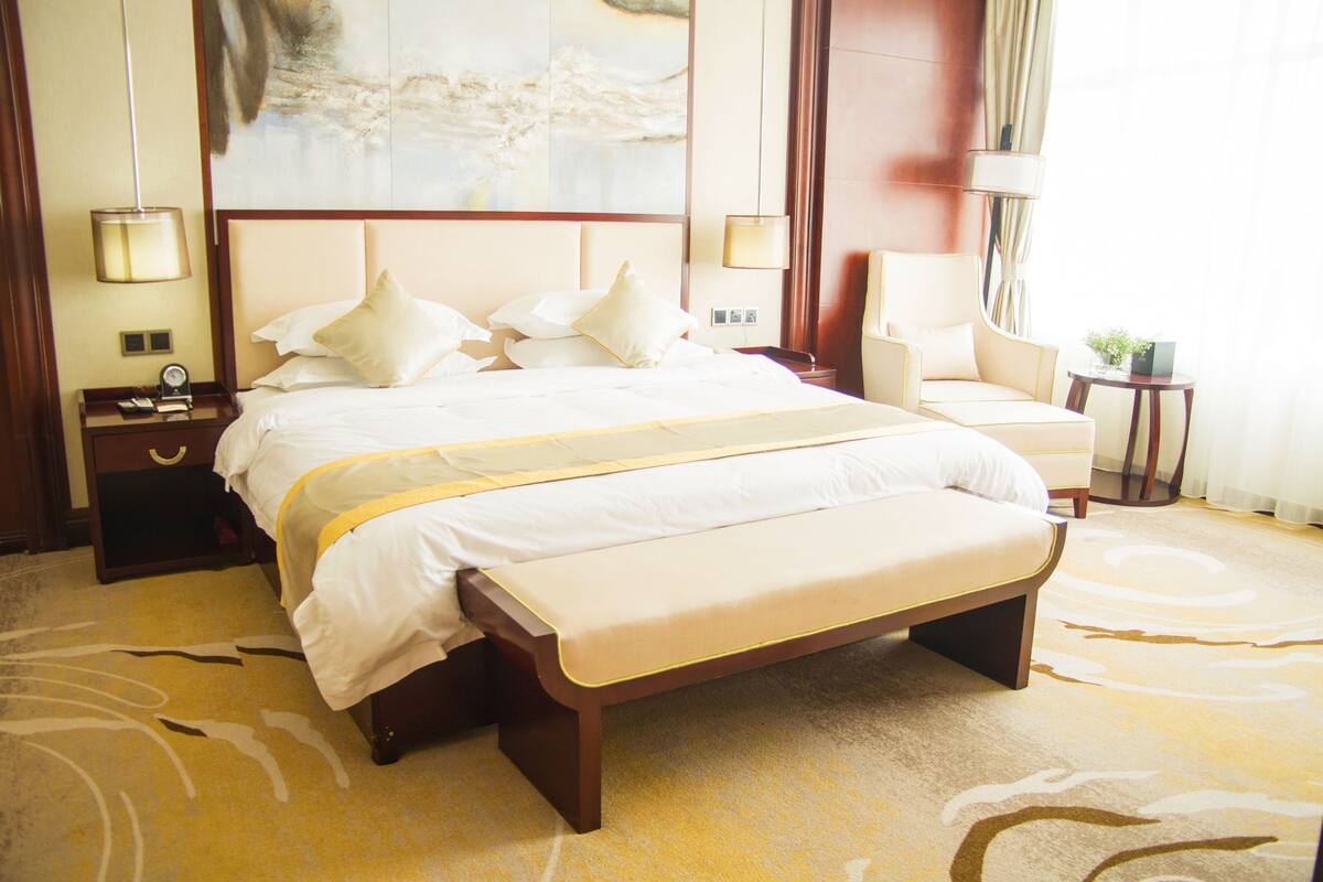 A large bed with a bench, a bedside table, a chair, and a small table with a plant.