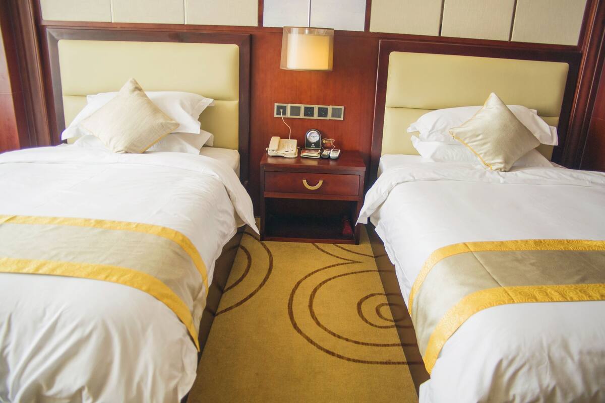 A hotel room with two beds, each with white bedding and a yellow accent. There is a wooden nightstand with a lamp, a phone, and a clock between the beds.