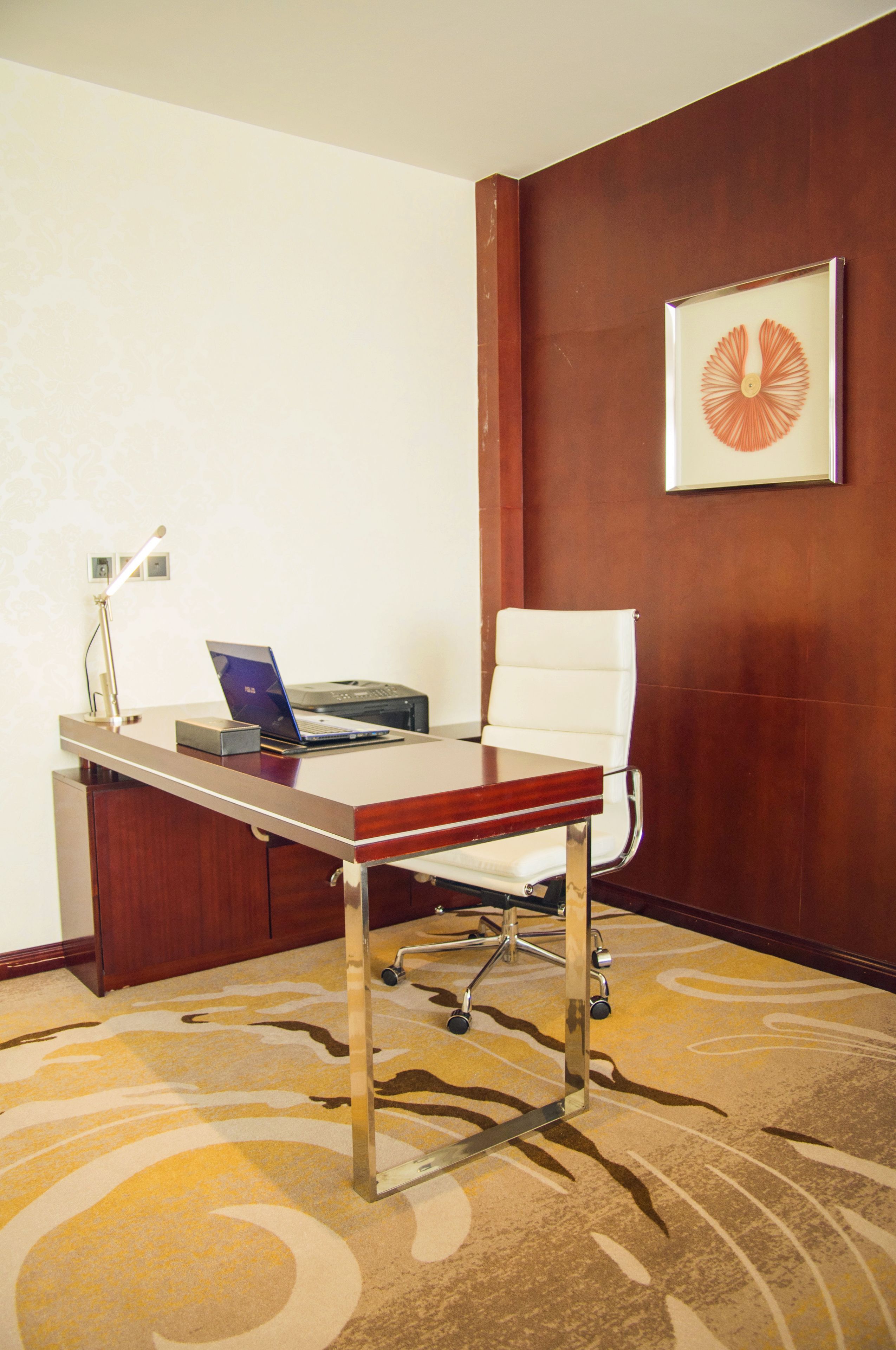 A hotel room with a desk, laptop, chair, and a framed picture on the wall.