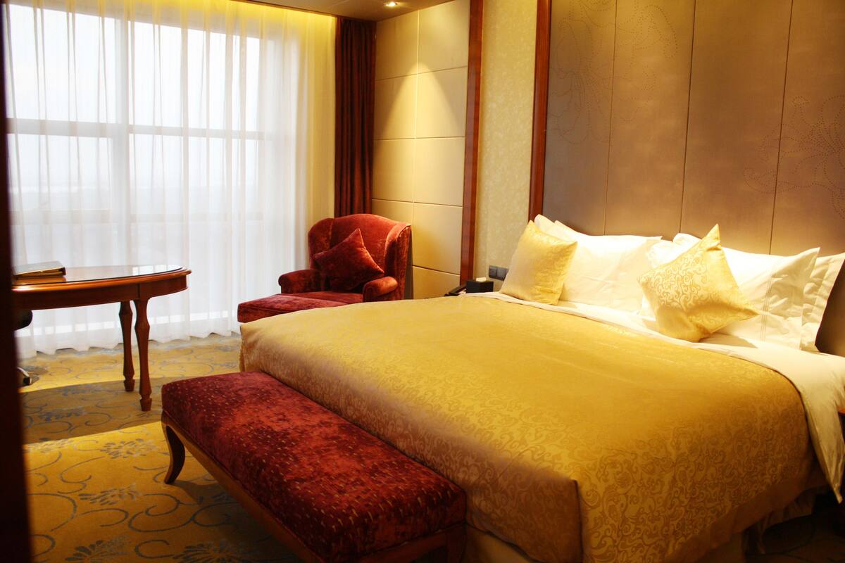 A hotel room with a large bed, a red armchair, a wooden nightstand, and a bench.
