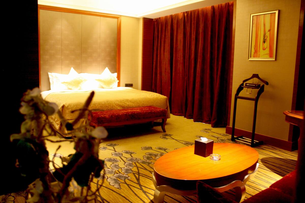 A hotel room with a bed, a bedside table, a chair, a small table, and a standing lamp.