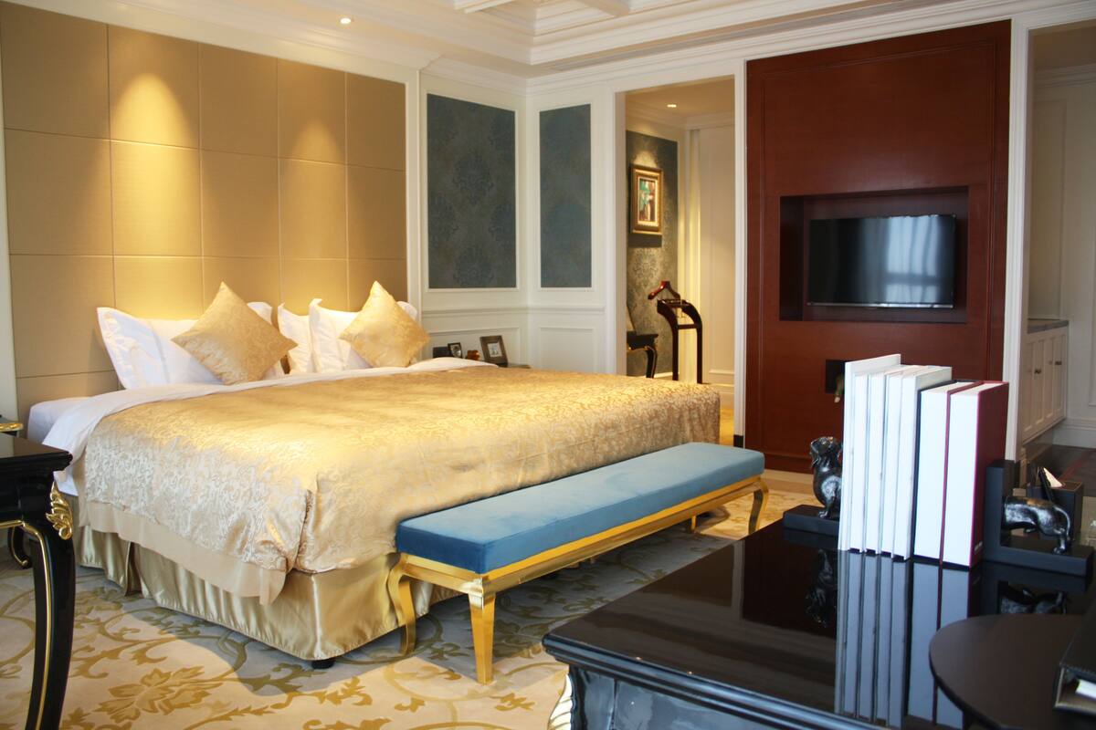 A large bed with a gold bedspread, a blue bench, and a TV mounted on a wooden panel.