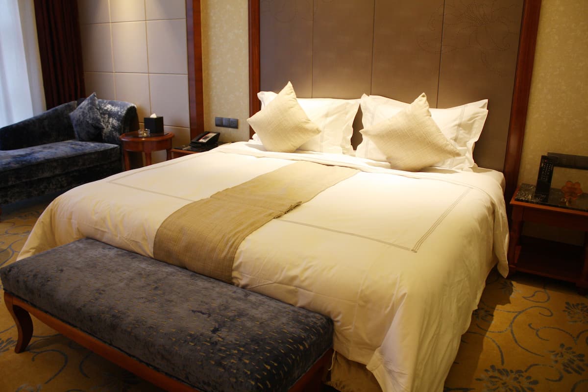 A neatly made bed with white linens and a beige throw, a dark upholstered bench, and a small bedside table with a lamp.