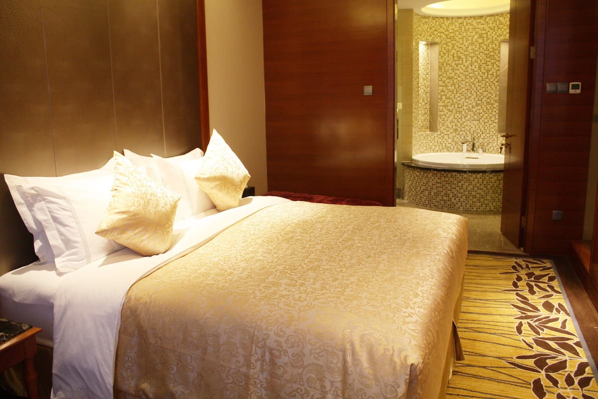 A hotel room with a large bed, a bedside table, a bathroom with a sink and mirror, and a carpeted floor.
