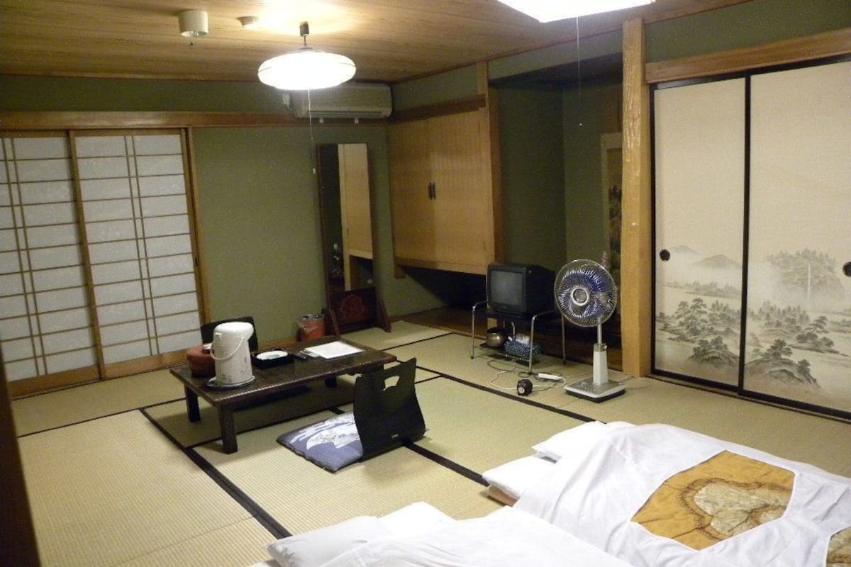 A traditional Japanese room with tatami flooring, a futon bed, a television, a fan, and a shoji screen.