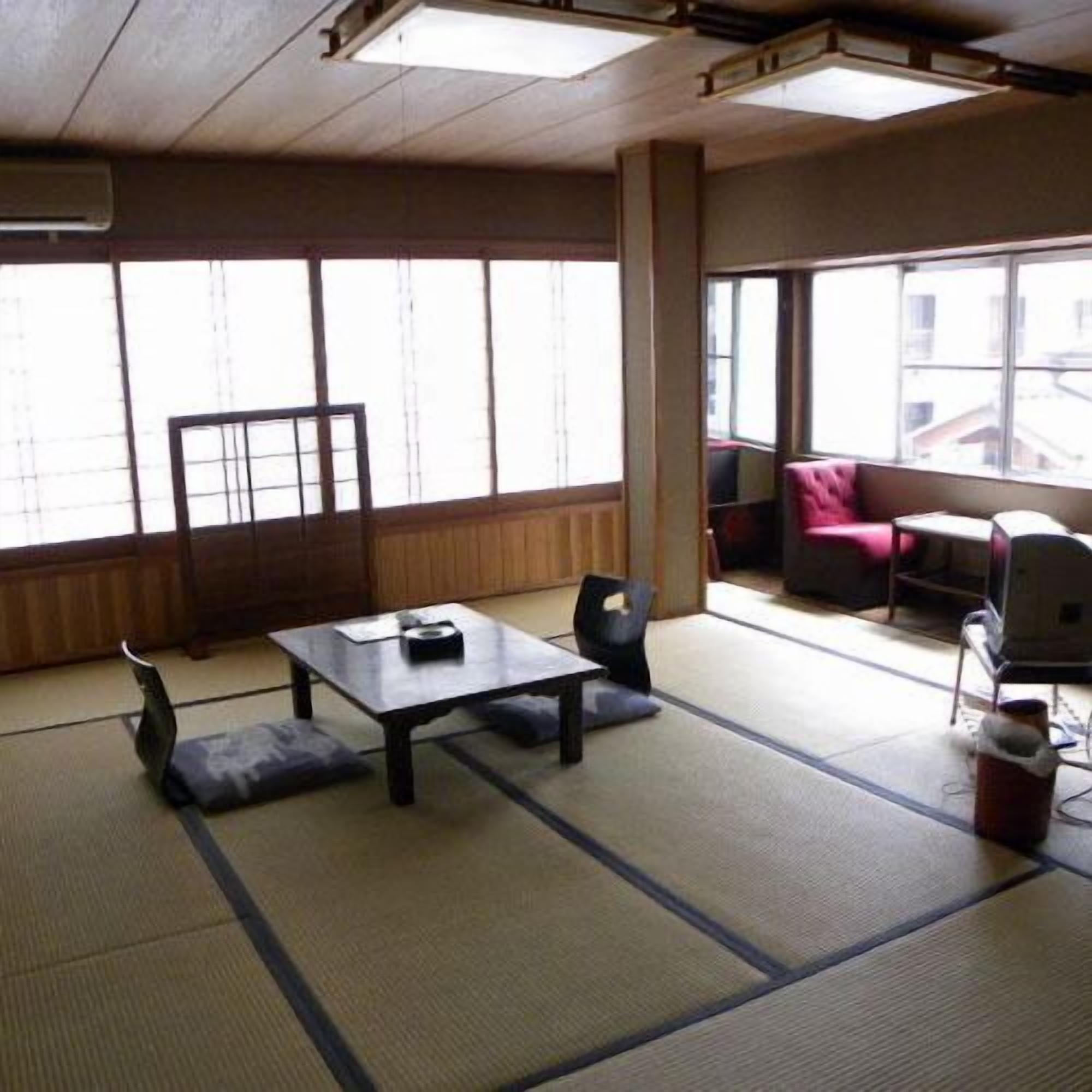 Ein Raum mit Tatami-Matten, einem niedrigen Tisch und Stühlen, großen Fenstern und Blick auf Gebäude.