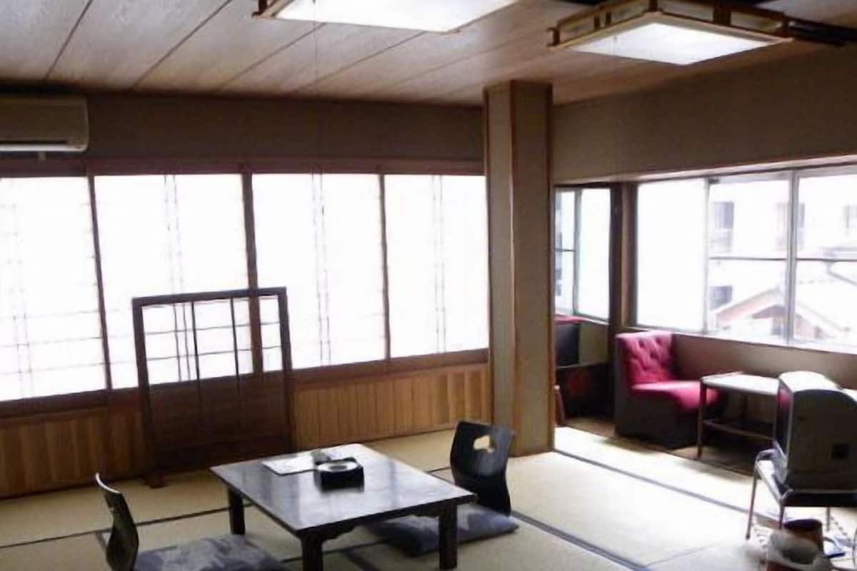 A tatami mat room with a low table and chairs, large windows, and a view of buildings outside.