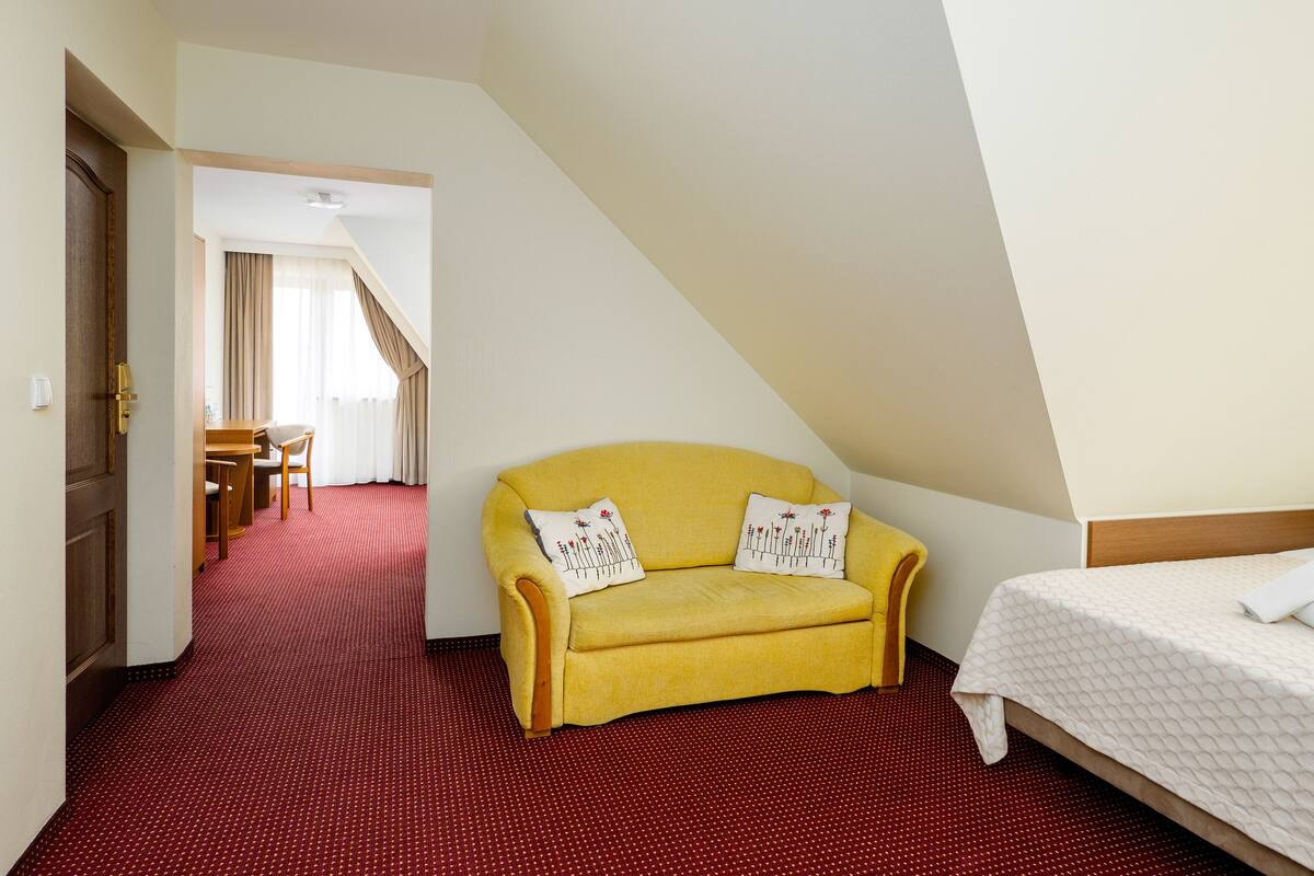 Habitación de hotel con un sofá amarillo, una cama, una alfombra roja, una puerta de madera y una zona de comedor con mesa y sillas.