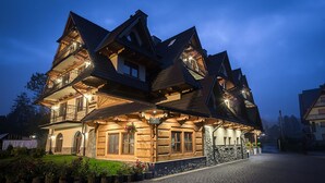 A large, multi-story wooden house with balconies and a stone foundation, surrounded by a cobblestone driveway.