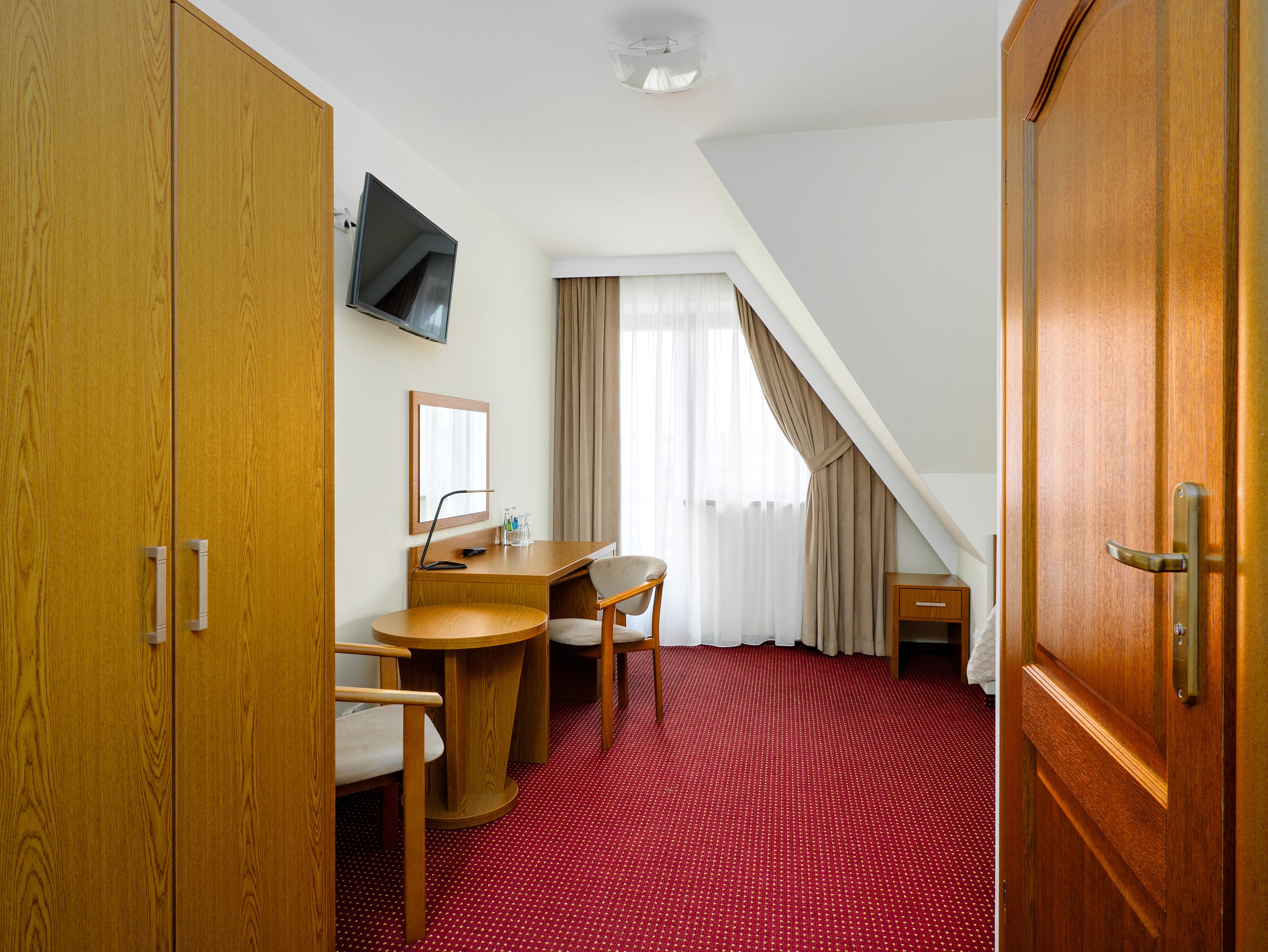 A hotel room with a wooden desk, a chair, a television mounted on the wall, and a large wooden door.