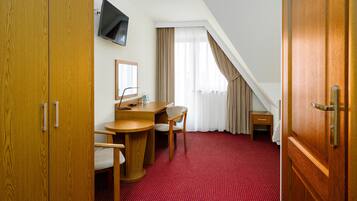 A hotel room with a wooden desk, a chair, a television mounted on the wall, and a large wooden door.