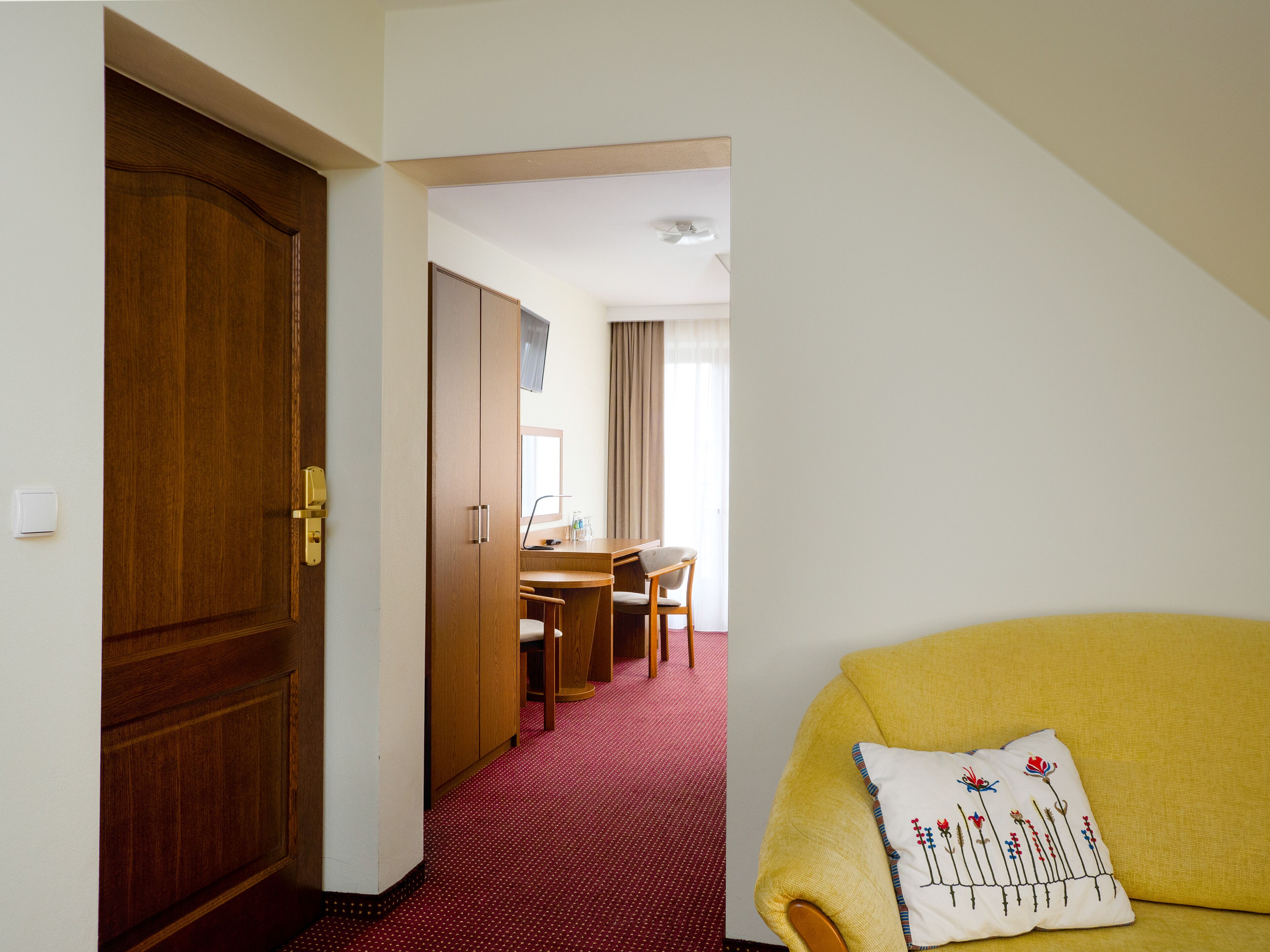 A hotel room with a wooden door, a desk with a chair, a television, and a yellow sofa with a decorative pillow.
