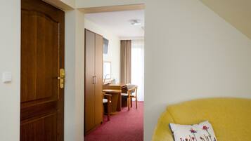 A hotel room with a wooden door, a desk with a chair, a television, and a yellow sofa with a decorative pillow.