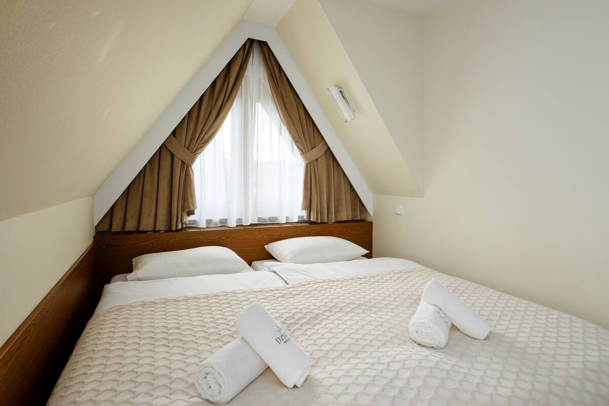 Un dormitorio con techo inclinado, cama con ropa blanca y ventana con cortinas beige.