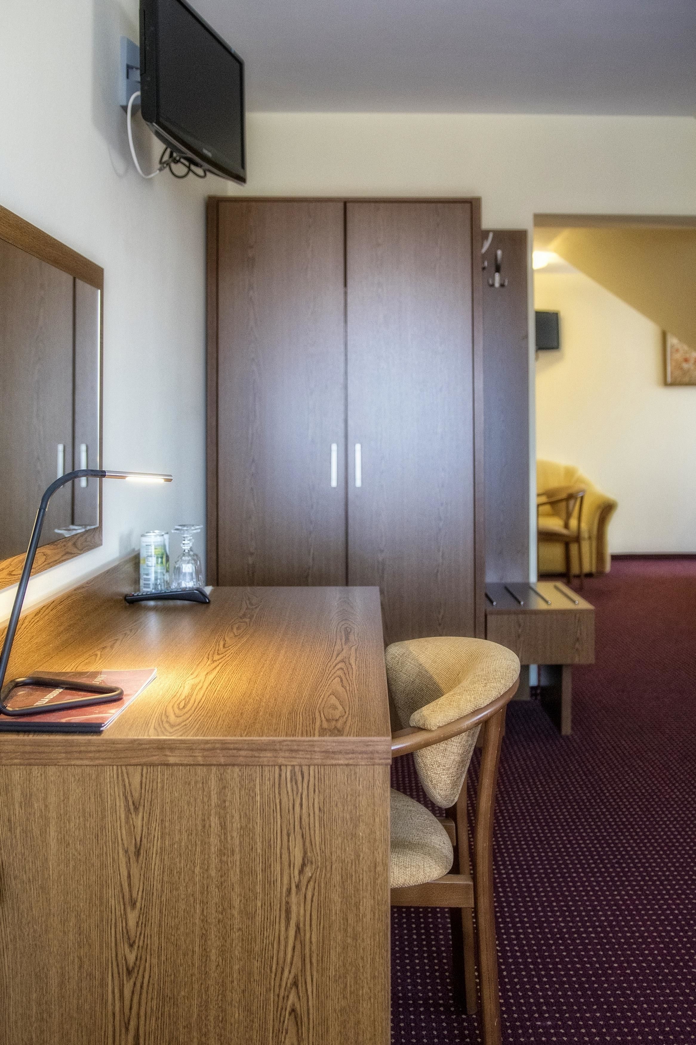 A hotel room with a wooden desk, a chair, a television mounted on the wall, and a wardrobe.